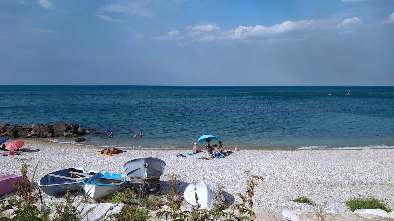 The photo depicts a white, medium-small pebble beach bordered by calm blue sea and clear sky. On the beach, there are blue and white wooden boats of the type used by fishermen. In the center of the photo, on the beach, is an beach umbrella with three people sitting underneath on beach chairs.