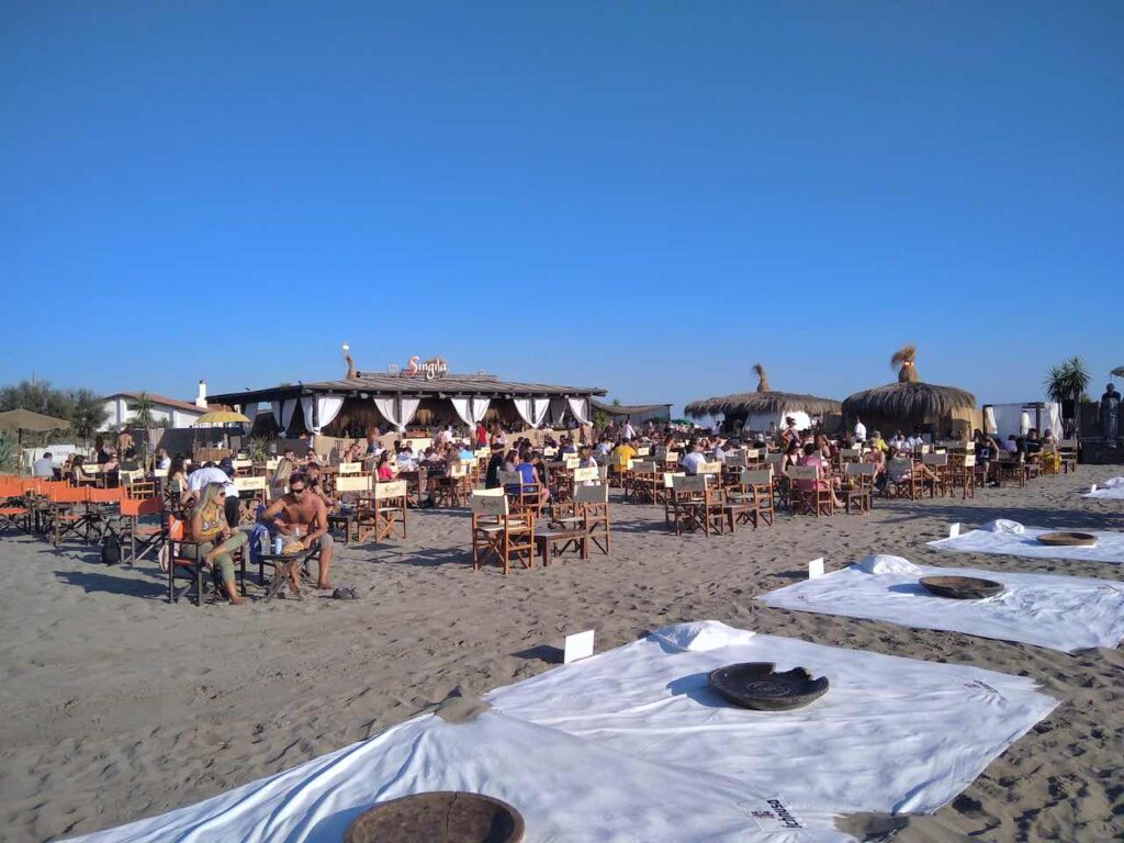 A gray sandy beach with wooden tables and stretched fabrics for those who want to have a drink sitting on the sand. There are people sitting at tables and behind them, a kiosk with white curtains.