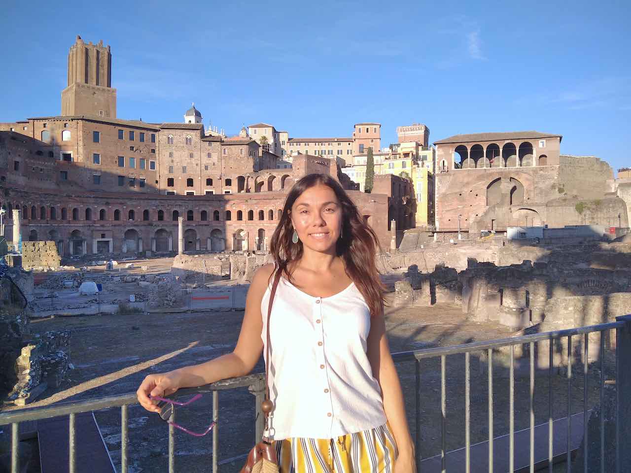 A young woman standing along Via dei Fori Imperiali in Rome, Italy. She's wearing a white top and light yellow trousers, the summer blue sky behind her. On the background, the profile of the Im perial Forum with the old brown-bricks buildings. she's smiling at the camera, happy to be there.