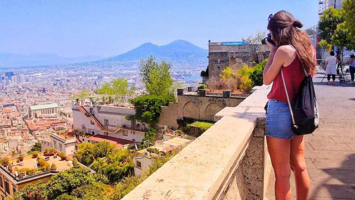 A tall girl with a camera in her hands and long brown hair is focused on taking a photograph of the view of Naples' historic center from the viewpoint in Montesano. In the distance, you can see the profile of the Vesuvius, mixing into the blue of the sea.