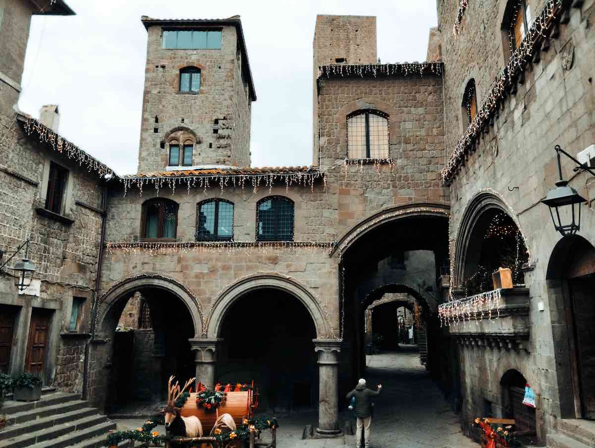 The photograph depicts a glimpse of a neighborhood with medieval architecture. The two-story houses are made of dark gray stone with mud-yellow hues. On the ground floor are arcades supported by slender columns, and slanted exterior stairways lead to the second floor. The houses are decorated with Christmas lights, and a wood sleigh can be glimpsed. The sky is cloudy.