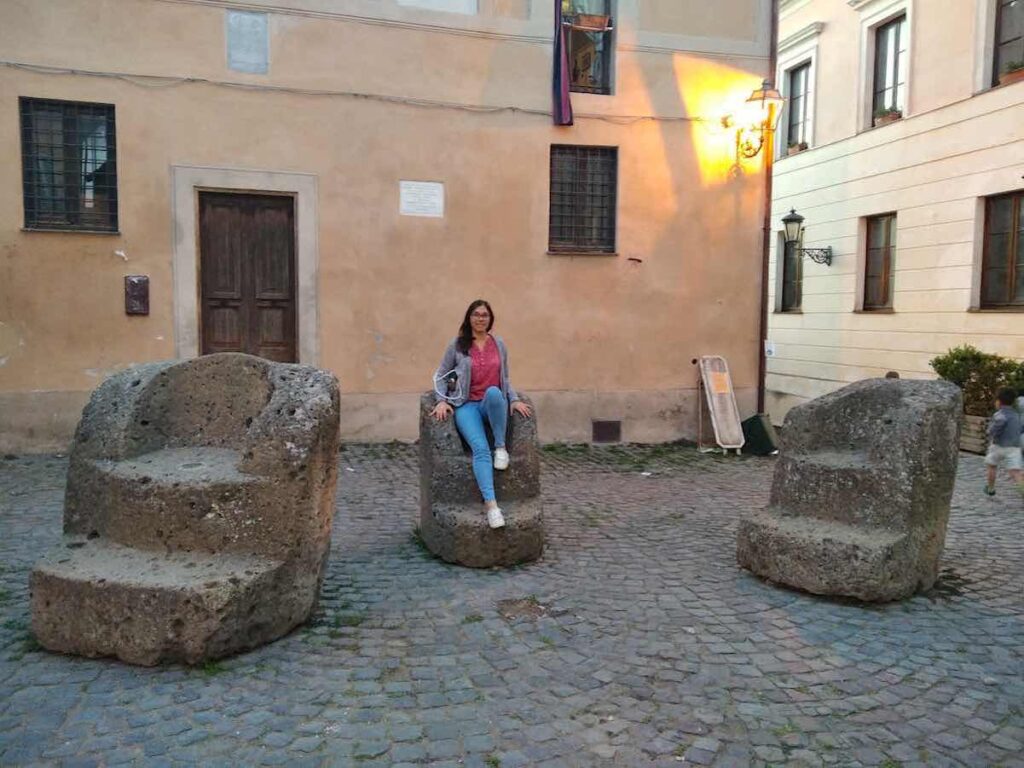 In a small square with a floor of sanpietrini, in a quaint small town near Rome, are three archaic-looking carved thrones, all of different sizes. On the central one sits a young woman with straight, shoulder-length brown hair and glasses. The woman wears jeans and a red shirt with white dots and smiles, satisfied.