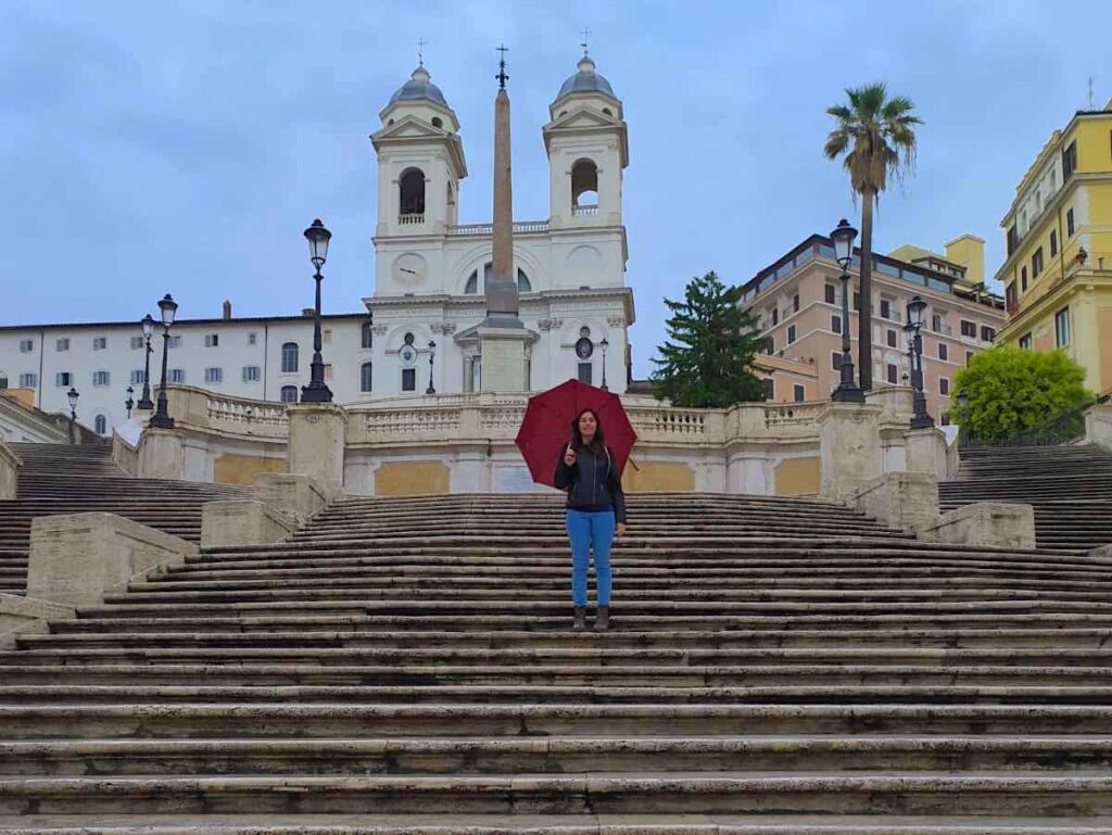 A young woman with long, straight brown hair, wearing a black jacket and a pair of jeans, stands holding a red umbrella in the middle of a large, long marble staircase. Behind the woman, there is the façade of an ancient church with twin steeples and, between the woman and the church, at the top of the steps, an Egyptian obelisk.