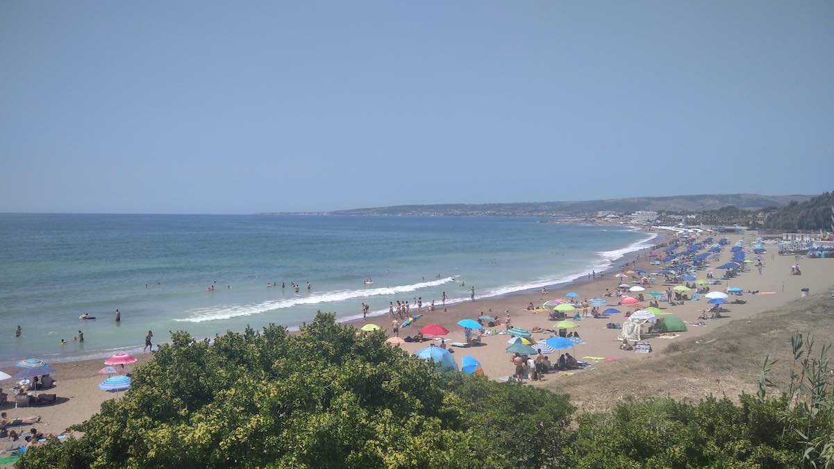 The Santa Severa beach near Rome offers a panoramic view of the Tyrrhenian Sea, lined with a vibrant array of beach umbrellas. Crowds of beachgoers enjoy the sun and surf, with the lush greenery in the foreground offering a natural contrast. This popular summer destination captures the essence of Italian coastal life, bustling with activity and scenic views.