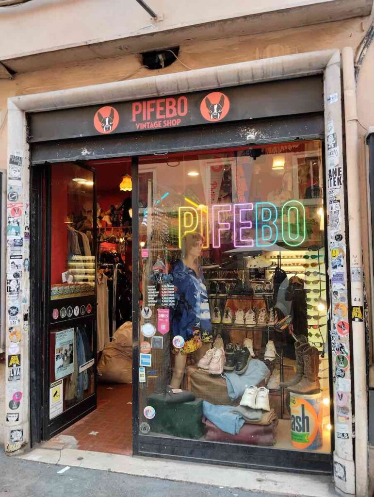 Exterior view of PIFEBO Vintage Shop in Rome's Monti neighborhood, showcasing a colorful storefront adorned with vibrant neon signs, eclectic stickers, and a display window inviting passersby to explore its retro fashion collection.