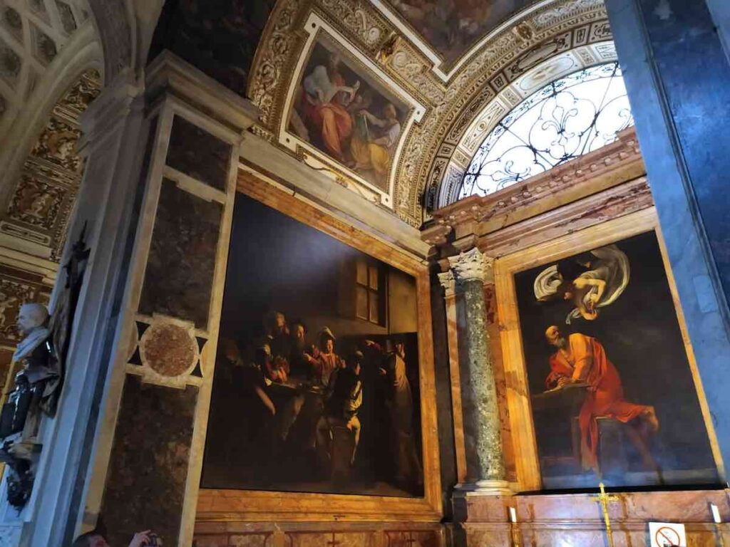 Interior of a richly decorated church featuring Caravaggio's paintings, 'The Calling of Saint Matthew' and 'The Martyrdom of Saint Matthew,' with intricate marble columns and an ornate ceiling