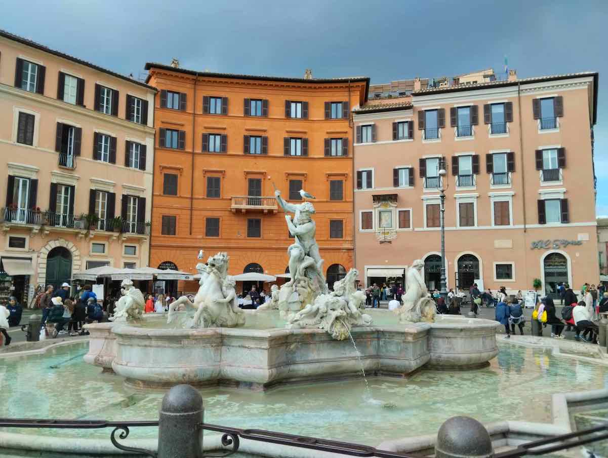 The Fountain of Neptune, featuring the sea god Neptune battling an octopus, surrounded by other mythical figures, set against vibrant orange buildings in the bustling Piazza Navona, Rome.