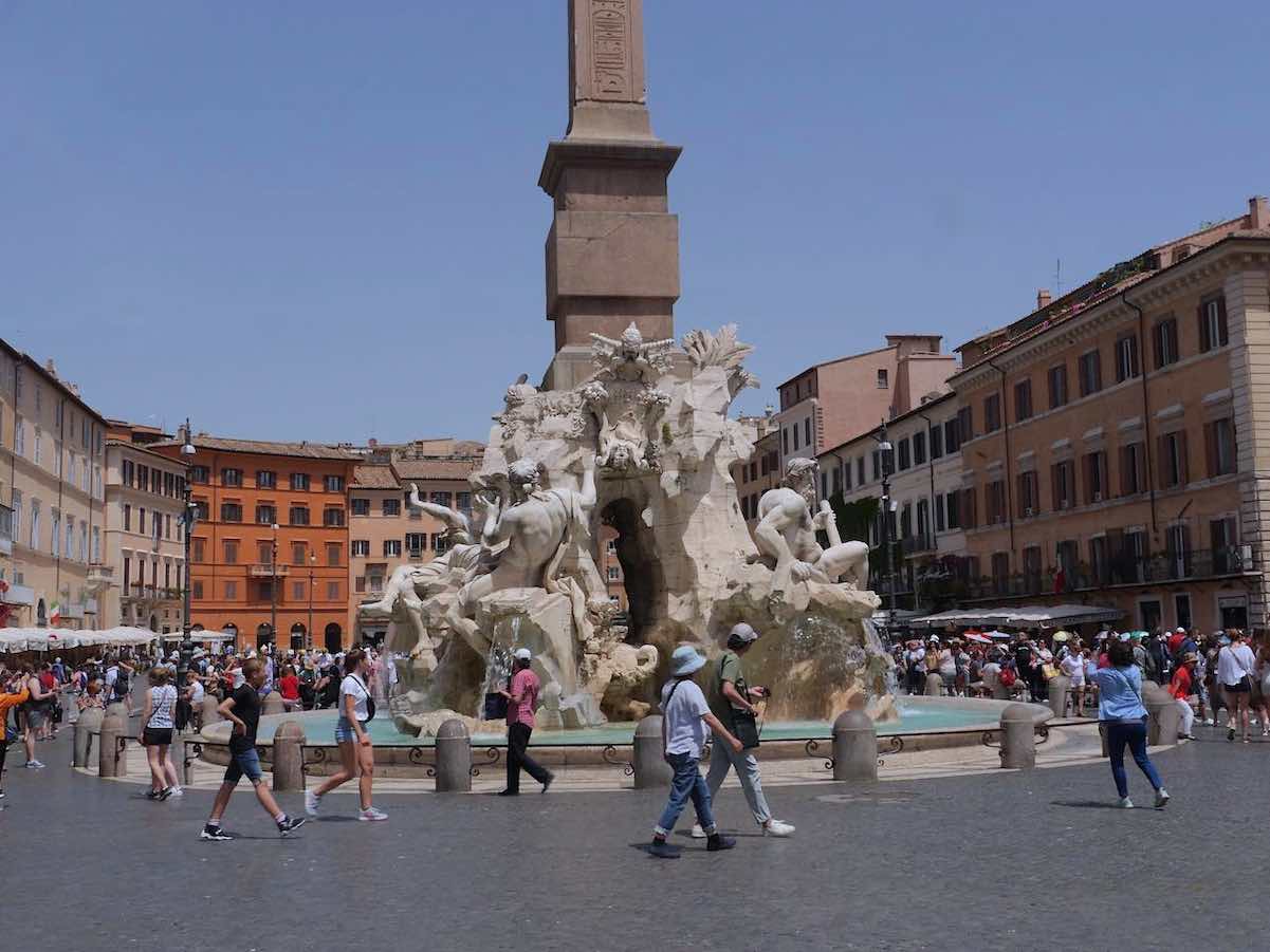 Tourists walking around the Fontana dei Quattro Fiumi, the Four Rivers Fountain, in Piazza Navona, Rome, with its impressive obelisk and sculpted figures representing major rivers, under a clear blue sky.