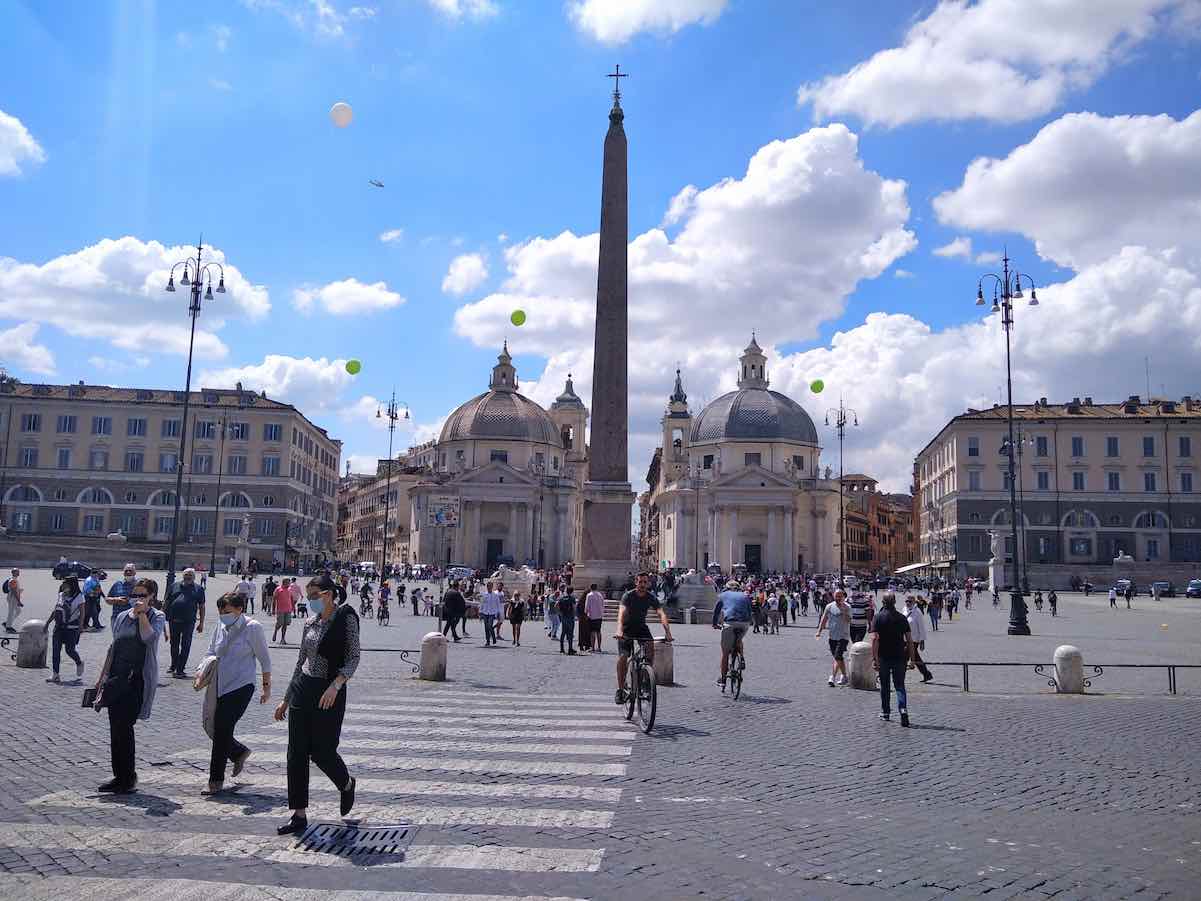 The image presents a vibrant scene of Piazza del Popolo in Rome. It's a sunny day with a bright blue sky adorned with fluffy clouds. The square is bustling with activity: pedestrians are walking across the cobblestone surface, some are engaged in conversations, while others appear to be in transit, heading to various destinations. A couple of cyclists are also navigating through the crowd.