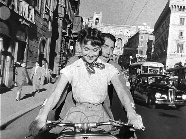 This iconic scene from "Roman Holiday" features Audrey Hepburn and Gregory Peck riding a Vespa through the bustling streets of Rome. Audrey Hepburn, dressed in a light-colored blouse and a playful neck scarf, sits in front, steering the scooter with a joyous smile. Gregory Peck sits closely behind her, holding on and enjoying the ride. The background showcases the vibrant city life of Rome with its historic architecture, vintage cars, and pedestrians, capturing the essence of the city's charm in the 1950s. The black-and-white imagery adds to the timeless allure of this classic film moment.