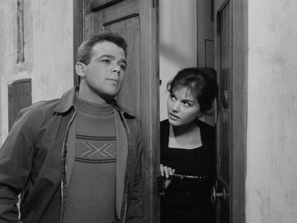 In this still from "Big Deal on Madonna Street," we see a young man, dressed in a patterned sweater and a jacket, standing confidently at the door. A woman peeks out from behind the partially opened door, secured with a chain, looking at him with a mix of curiosity and skepticism. The black-and-white cinematography adds a timeless, classic feel to the scene, reflecting the film's vintage charm.