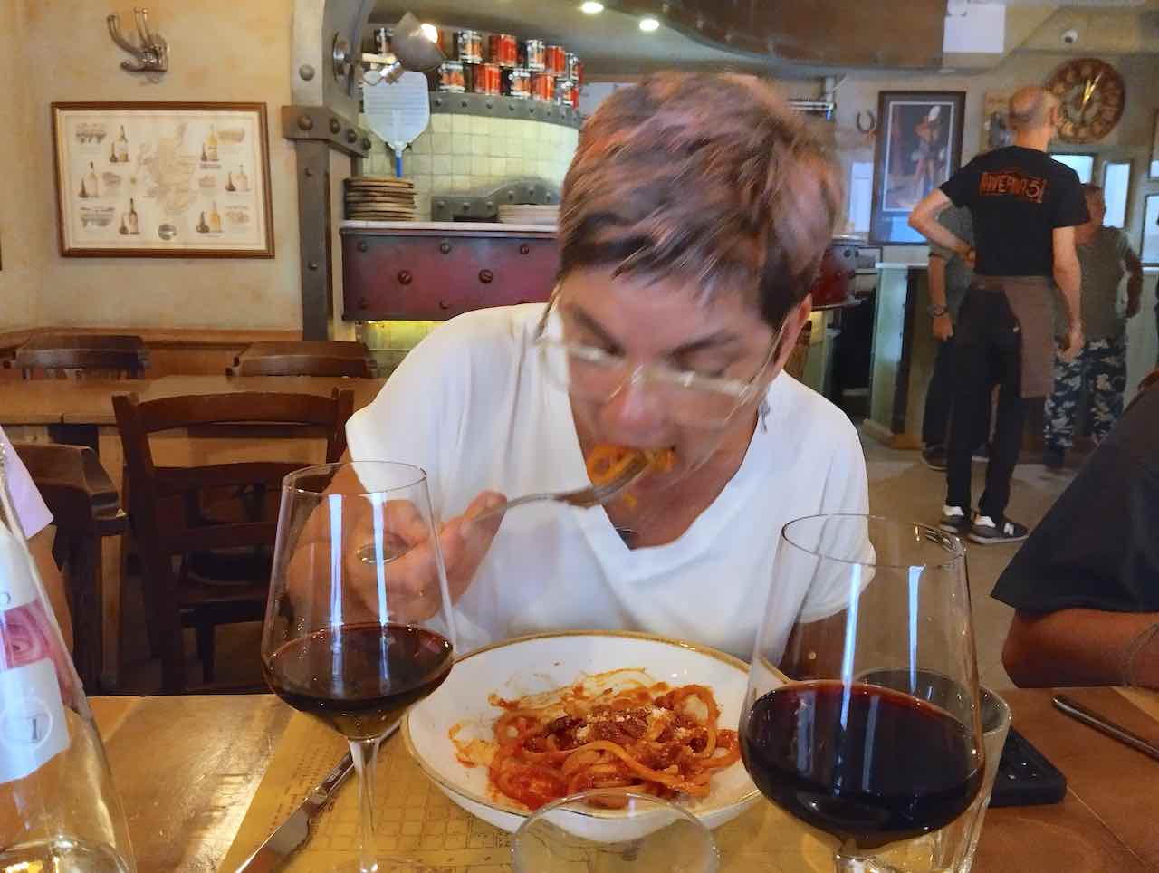 A woman is eating a hearty dish of pasta with tomato sauce sitting at a table in what looks like a Roman trattoria. In front of her are two glasses with red wine.