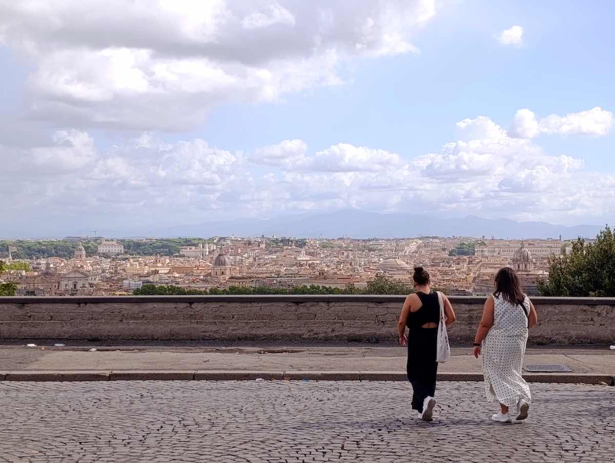 Two women in summer dresses are walking towards the edge of a large terrace on the Janiculum Hill, one of the panoramic hills of Rome.